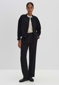 Zwarte jack met een ronde kraag en knoopsluiting, gecombineerd met zwarte op maat gemaakte broek. Glad textuur, ontspannen pasvorm en minimalistisch ontwerp.
