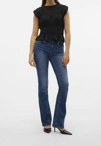 Svart smockad topp med volangkanter, parat med blå utsvängda jeans och svarta spetsiga klackar; slät tyg med en figurnära silhuett.