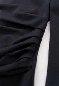 Schwarzer Stoff mit einer weichen Textur, der an einer Seite geraffte Details aufweist und einen drapierten Effekt gegen eine glatte Oberfläche erzeugt.