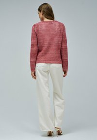 Rosa gestrickter Pullover mit offenem Gewebemuster, langen Ärmeln und rundem Ausschnitt, kombiniert mit weißen, weit geschnittenen Hosen.