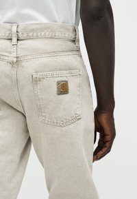 Πλάγια όψη ατόμου που φορά ανοιχτό μπεζ τζιν Carhartt με εμφανές λογότυπο στην πίσω τσέπη και σκούρο δέρμα στο δεξί χέρι που βρίσκεται χαλαρά δίπλα στο σώμα.