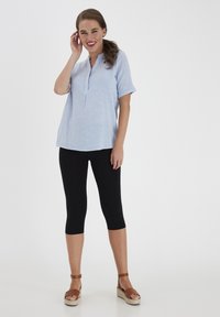 Jasnoniebieska koszula z krótkim rękawem z detalem przy guziku, zestawiona z czarnymi legginsami i brązowymi espadrylami. Gładka tekstura, luźny krój.