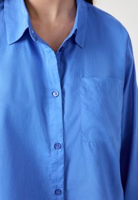 Niebieska koszula z kołnierzykiem, z naszywaną kieszonką po lewej stronie. Tkanina ma gładką teksturę i delikatny połysk.