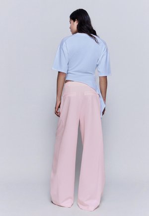 Femeie cu păr lung și închis la culoare, purtând un top albastru deschis cu mâneci scurte și pantaloni largi de culoare roz pal, cu talie înaltă, văzută din spate, pe fundal simplu.