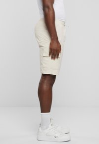 Beige cargo-shorts med sidelommer, laget av lett materiale, iført hvite sneakers og hvite sokker, vist fra en sidevinkel.
