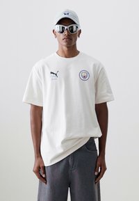 Bílý bavlněný tričko s krátkými rukávy, se znakem Puma na levé hrudi a kulatým odznakem Manchester City. Nosit s šedými šortkami a slunečními brýlemi.