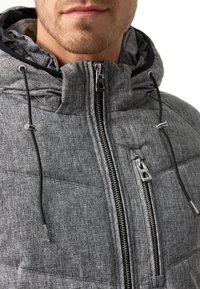 Mies, ktorý má na sebe šedú prešívanú bundu s kapucňou, čierne šnúrky a dva viditeľné strieborné zipsy na bielom pozadí.