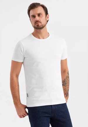 Weißes Kurzarm-T-Shirt mit Rundhalsausschnitt und glatter Textur. Kombiniert mit dunklen Jeans, die ein Tattoo am Arm zeigen.