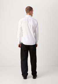 Hvit langermet skjorte med knappdetaljer, sammen med svarte bukser, som viser et enkelt design og en figursydd form fra bakvinduet.