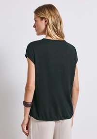 Top nero a maniche corte con scollatura rotonda e orlo posteriore leggermente arricciato, realizzato in un tessuto morbido. Indossato con pantaloni di colore chiaro.