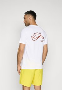 Hvit T-skjorte med rød "Giddy Up" grafikk på ryggen, sammen med gule shorts. Mykt stoff med rund hals og korte ermer.