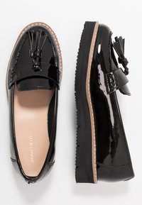 Sorte patent lær loafers har en dekorativ dusk, en rund tå og en kontrasterende teksturert såle med sydd detaljering.