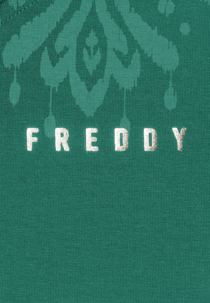 Un tessuto verde con una texture a coste presenta la parola "FREDDY" in lettere metalliche argentate, accompagnata da un intricato motivo sbiadito.