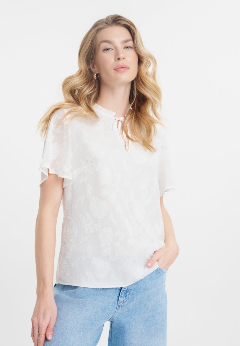 Blusa blanca de manga corta con bordado floral, escote redondeado y un pequeño detalle de lazo. Tejido suave y transparente sobre un corte holgado.