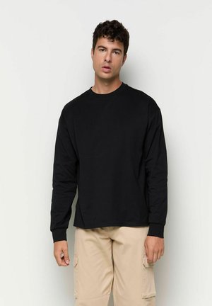 Junger Mann mit lockigem dunklem Haar, der ein schwarzes Langarmshirt und beige Cargohosen trägt, steht vor einem schlichten hellen Hintergrund.