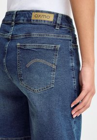 Prim-plan cu o persoană care poartă blugi albaștri din denim, cu un buzunar posterior cusut și o etichetă bej „oxmo” pe talie.
