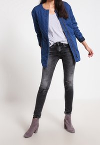 Blå quiltad jacka, grå randig t-shirt, svarta slitna skinny jeans och grå ankelstövlar med blockklack på en ljus bakgrund.