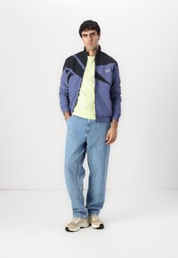 Letvægts blå jakke med sorte detaljer over en gul t-shirt, parret med afslappede denim jeans og beige sportsko.