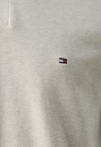 Helles beige Polohemd aus strukturiertem Stoff mit einem kleinen Logo in Rot, Weiß und Blau auf der linken Brustseite.