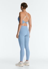 Woman stands facing away wearing light blue leggings and a matching blue sports bra with white crossed straps.