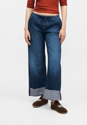 Άτομο που φοράει φαρδιά μπλε τζιν με γυρισμένα μανίκια, καφέ σουέντ αθλητικά παπούτσια και κόκκινο μακρυμάνικο τοπ, στέκεται μπροστά σε απλό φόντο.