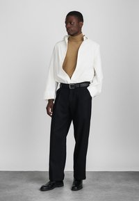 Camisa oversized branca com colarinho de xale, gola alta bege por baixo, calças plissadas pretas e sapatos de couro pretos. Fundo simples.