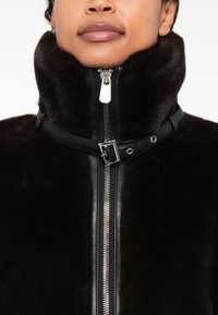 Casaco de pele preto com um colarinho alto e um fecho metálico de comprimento total com acabamentos em pele. Textura suave com detalhes notáveis.