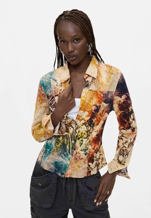 Woman with braided hair wears a multicolored abstract print button-up shirt over a white top and dark cargo pants with large pockets.