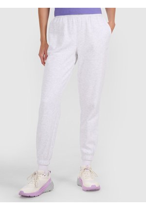Persona con pantalones deportivos gris claro, camiseta morada, calcetines blancos y zapatillas crema con suelas y detalles en lavanda, de pie con las manos en los bolsillos.