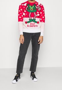 Persona que lleva un suéter navideño rojo y blanco con un diseño de elfo y el texto "Elf & Safety", jeans negros y zapatillas en blanco y negro.