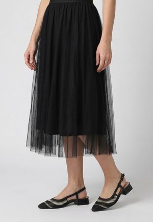 Kobieta ubrana w czarną tiulową spódnicę midi i czarne buty typu slingback na niskim obcasie z beżowym plecionym wzorem.