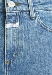 Primer plano de unos vaqueros de mezclilla azules que muestran un botón de metal y una etiqueta con la estampilla "CLOSED" cerca del bolsillo delantero y el lazo del cinturón.