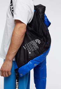 Niebieska kurtka puchowa z błyszczącym wykończeniem, czarną podszewką z tekstem, zamkiem błyskawicznym i zatrzaskami. Posiada pasek do noszenia.