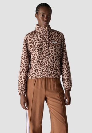 Woman wearing a leopard print zip-up jacket with high collar and brown pants with side stripes, standing against a plain light background.