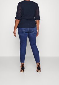 Marineblaues Oberteil mit durchsichtigen Polka-Dot-Ärmeln, kombiniert mit eng anliegenden dunkelblauen Jeans und schwarzen High Heels. Rückansicht, die Passform und Design präsentiert.