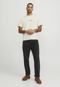 Beige Baumwoll-T-Shirt mit kurzen Ärmeln, auf dem gedruckter Text zu sehen ist, kombiniert mit schwarzen Hosen und braunen Schnürstiefeln. Einheitlicher Hintergrund.