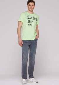 Camiseta de manga corta verde claro con texto impreso en negro, combinada con jeans grises y zapatillas blancas. La tela parece suave y casual.