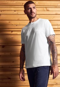 Muž s tetovanými pažemi, oblečený v bílém tričku s nápisem "Crafted Goods" a tmavých kalhotách, stojí proti dřevěné zástěně a dívá se nahoru.