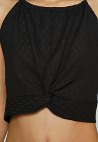 Crop top noir texturé sans manches avec un nœud torsadé à l'avant, porté par une personne avec un ventre visible et un teint clair.