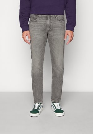 Mann trägt ausgewaschene graue Slim-Fit-Jeans, grün-weiße Sneakers und ein dunkelviolettes Langarmshirt, steht vor einem einfarbigen Hintergrund.