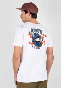 T-shirt en coton blanc affichant un graphique bleu d'un cochon portant un casque avec des ailes, agrémenté de touches de rouge et d'orange, et du texte qui indique "OXBOW STAY ALIVE."