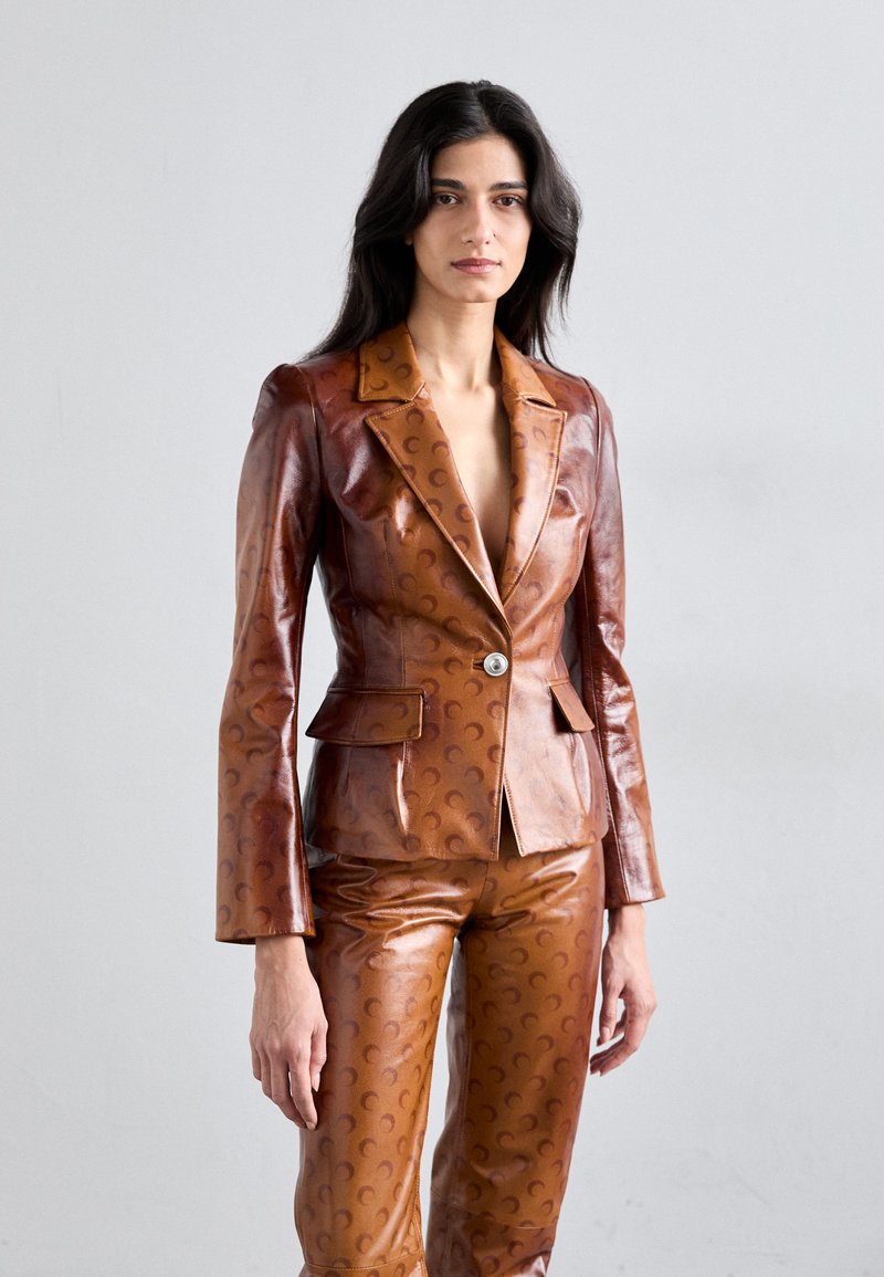 Costum din piele maro, cu un sacou ajustat și pantaloni, având un model subtil în formă de semilună, rever cu tăietură în formă de V și închidere cu un singur buton.