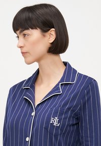 Camisa de pyjama azul marinho riscada com detalhes em branco, com fechamento de botão na frente e um bolso no peito esquerdo que exibe um logotipo bordado "RL". Textura de tecido suave.
