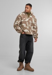 Pullover in pile marrone e crema con zip a metà, vestibilità oversize e motivo testurizzato; abbinato a jeans neri e stivali in pelle color cammello con lacci.