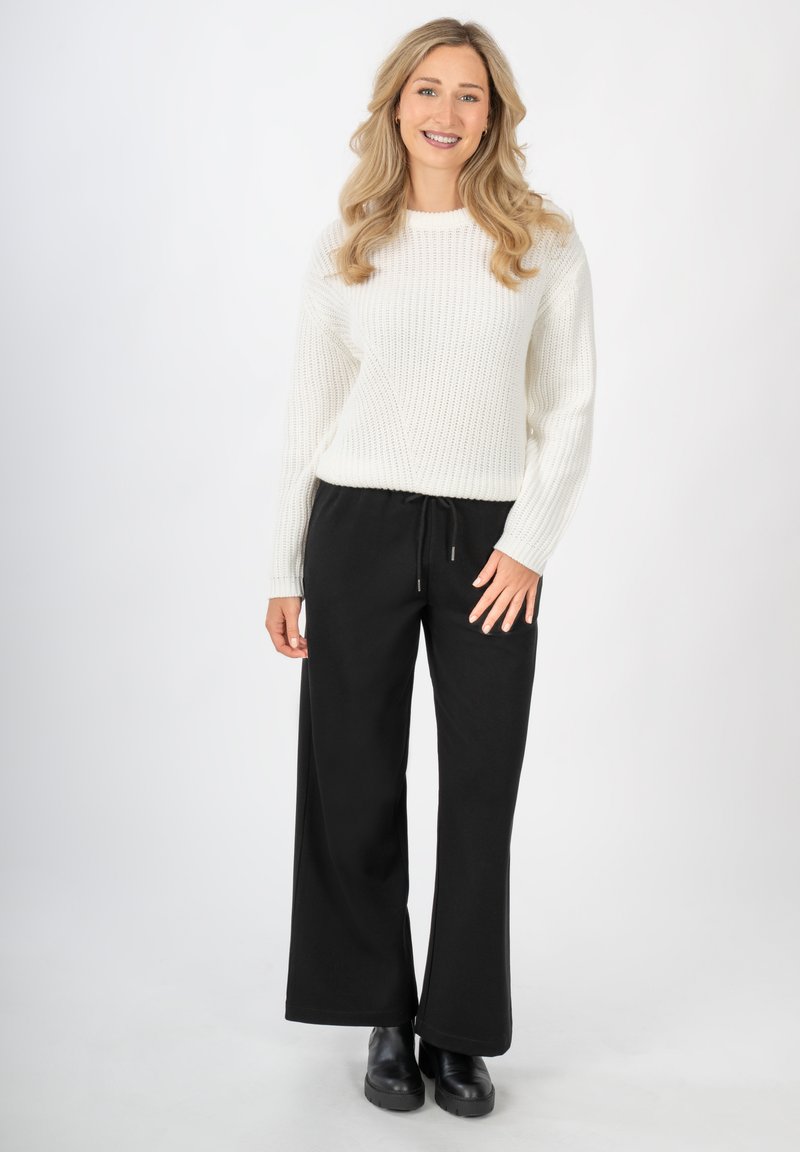 Donna sorridente con lunghi capelli biondi, che indossa un maglione bianco lavorato a maglia, pantaloni neri a gamba larga e stivali neri, in piedi davanti a uno sfondo semplice.
