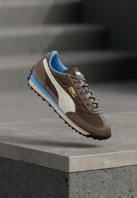 Braune und blaue Puma Easy Rider Sneaker, die über einer Betonkante schweben, mit einem verschwommenen grauen Treppenhintergrund.