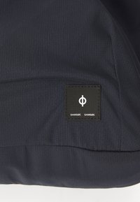 Navy fabric jacket featuring a textured surface, with a black rectangular logo patch displaying stylized white lettering and symbol.
