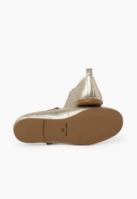 Scarpe piatte color oro metallico con punta arrotondata, tacco piccolo e dettaglio con cinturino. La suola è marrone con la taglia della scarpa e il paese di origine stampati.