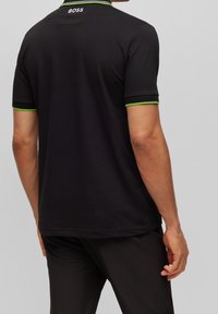 Svart polo t-shirt med grön kant, med 'BOSS'-logotyp på baksidan av halsringningen. Korta ärmar och klassisk kragdesign. Slät textur.