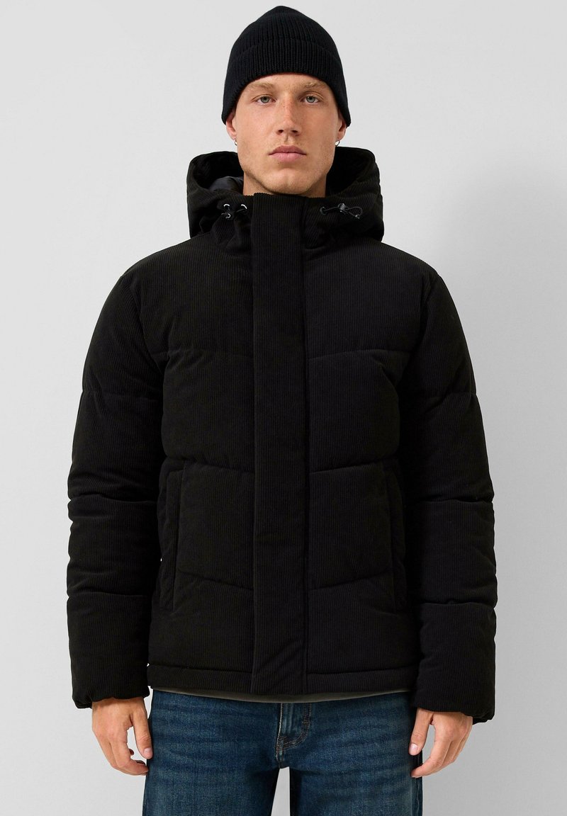 Giacca puffer in corduroy nera con cappuccio, dotata di zip frontale, polsini elastici e due ampie tasche frontali. Indossata con un berretto nero.